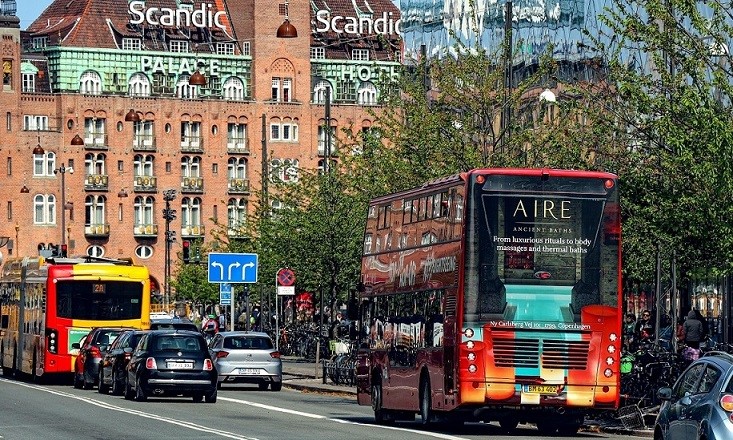 Busreklame for AIRE Ancient Baths på en rød dobbeltdækkerbus i Københavns bymiljø, med Scandic Hotel i baggrunden, illustrerer effektiv reklameplacering i en travl by.